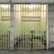 Row of small prison cells with bars in front. Inside the cell is a bed and toilet.