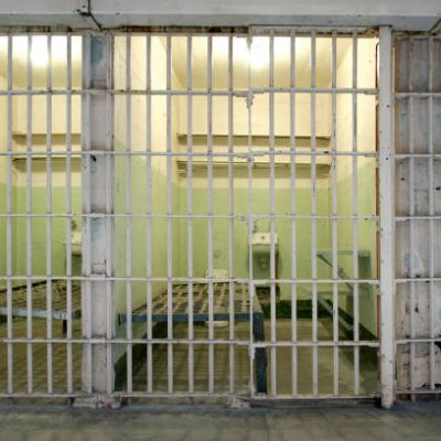 Row of small prison cells with bars in front. Inside the cell is a bed and toilet.